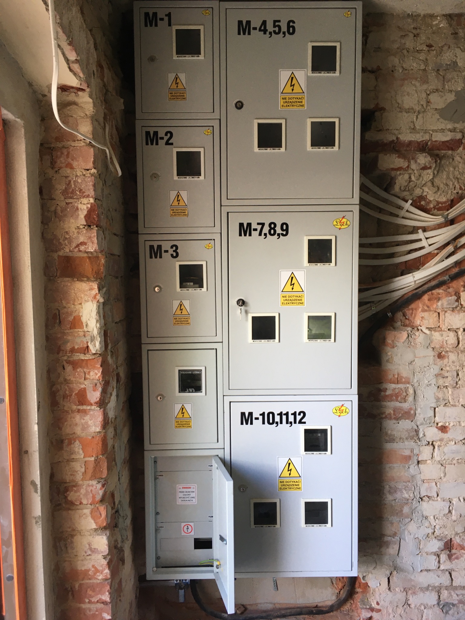 Rozdzielnica elektryczna z wieloma licznikami, zamontowana na ceglanej ścianie w surowym stanie, z widocznymi przewodami i otwartą jedną z szafek.