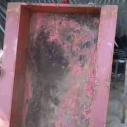 Czerwona, metalowa konstrukcja z dźwignią, częściowo oczyszczona z rdzy i starej farby, stojąca na piasku.