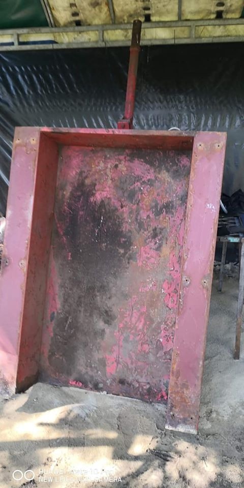 Czerwona, metalowa konstrukcja z dźwignią, częściowo oczyszczona z rdzy i starej farby, stojąca na piasku.