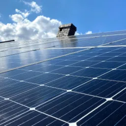 Panele słoneczne na dachu domu z ceglanym kominem w tle, widoczne odbicie nieba i chmur.