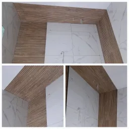 Wnętrze łazienki w trakcie wykańczania: białe płytki imitujące marmur na ścianach, drewniane panele lamelowe na suficie i fragmencie ściany, widoczne przewody instalacji.