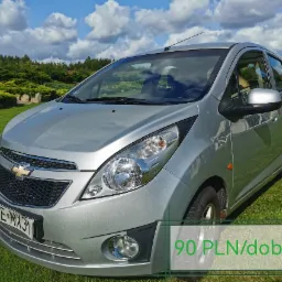 Srebrny Chevrolet Spark zaparkowany na trawie, z widoczną tablicą rejestracyjną GLE MX31 i informacją o cenie 90 PLN za dobę, na tle zieleni i błękitnego nieba z chmurami.
