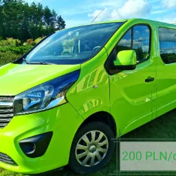 Żółto-zielony Opel Vivaro, widok z przodu i boku, zaparkowany na trawniku przed zielonymi drzewami i błękitnym niebem; na dole widoczny napis '200 PLN/doba'.
