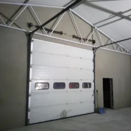Biała segmentowa brama garażowa z oknami, zamontowana w obiekcie we Władysławowie, ściany po świeżo wykonanych pracach tynkarskich, widoczna konstrukcja dachu.