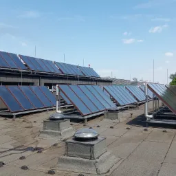 Rząd paneli słonecznych na płaskim dachu budynku w słoneczny dzień, widoczne r&oacute;wnież świetliki dachowe i elementy instalacji.