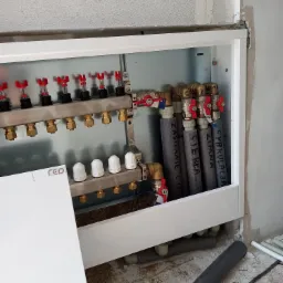 Rozdzielacz ogrzewania podłogowego w metalowej szafce, widoczne rury z izolacją i oznaczeniami 'Zasilanie C.O.', 'Ciepła', 'Zimna', 'Cyrkulacja', manometry z czerwonymi pokrętłami, klucz nastawny...