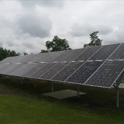 Rząd paneli słonecznych na metalowych wspornikach, zainstalowany na trawiastym terenie z betonową podstawą, widoczny szary osprzęt i ogrodzenie z betonowych bloczków w tle pod pochmurnym niebem.