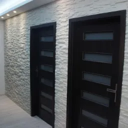 Korytarz z trzema ciemnymi drzwiami wewnętrznymi z przeszkleniami, wykończony dekoracyjną ścianą z białego kamienia i oświetleniem punktowym.