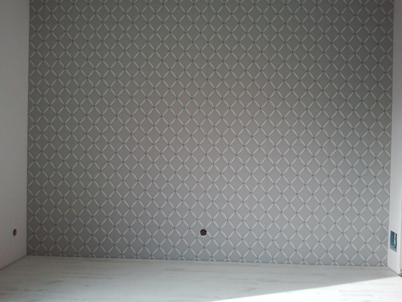Ściana wykończona tapetą w geometryczny wzór rombów z delikatnymi białymi akcentami, widoczny fragment podłogi w jasnym odcieniu szarości i fragment ściany bocznej z otworem na gniazdko.