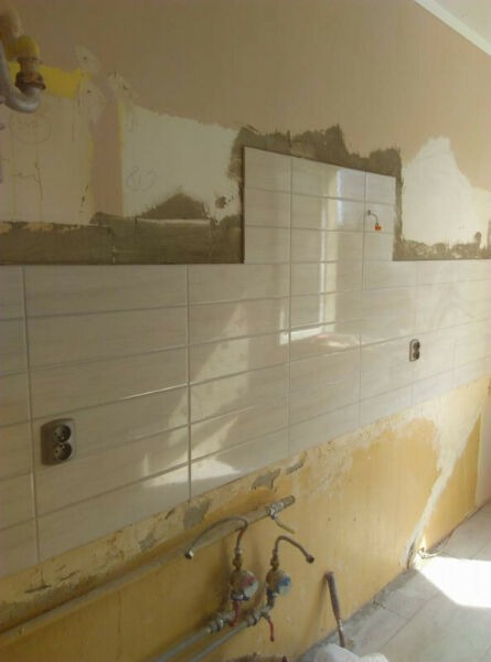 Ściana w trakcie układania płytek ceramicznych w łazience, widoczne fragmenty starej farby i instalacji hydraulicznej, gniazdko elektryczne na świeżo położonych płytkach.