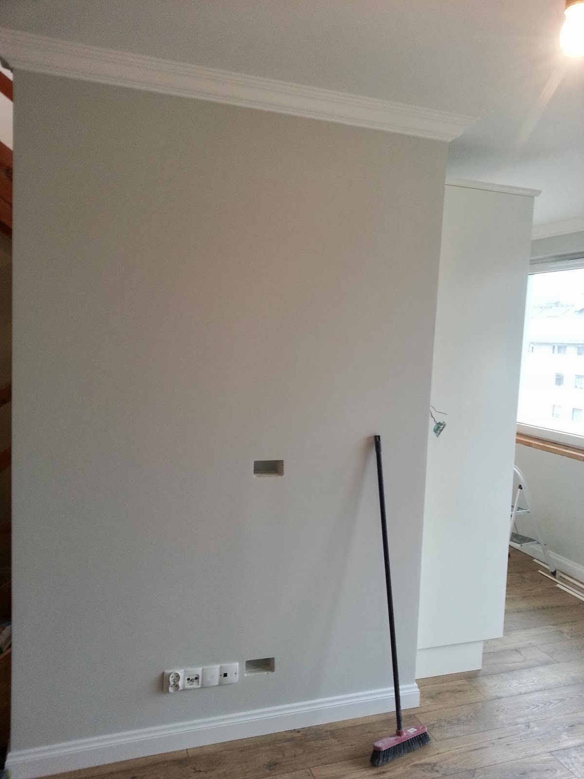 Świeżo pomalowana szara ściana z listwami przypodłogowymi i sufitowymi, gniazdkami elektrycznymi, otworami na kable i opartą o nią szczotką na drewnianej podłodze.