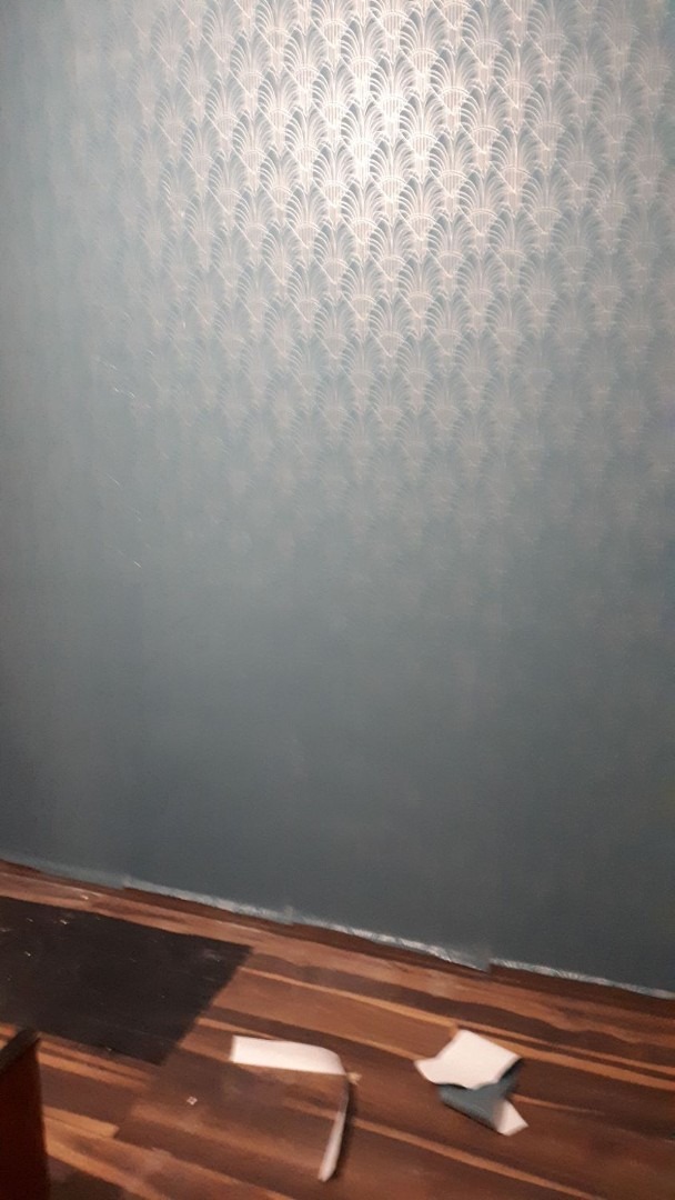 Ściana pomalowana na niebiesko-szaro z delikatnym wzorem w kształcie wachlarzy, widoczne niedociągnięcia przy podłodze, fragmenty podłogi z paneli i resztki materiałów budowlanych.