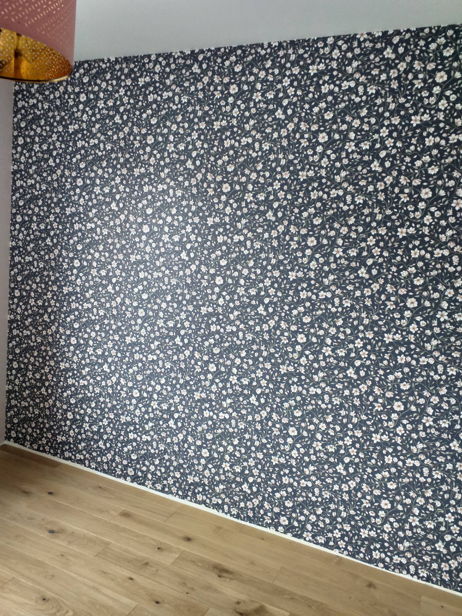 Ściana pokryta ciemną tapetą w drobne, białe kwiatki, panele podłogowe w odcieniach brązu, widoczny fragment lampy z ażurowym abażurem.