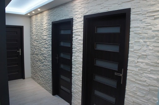 Korytarz z trzema ciemnymi drzwiami wewnętrznymi z przeszkleniami, wykończony dekoracyjną ścianą z białego kamienia i oświetleniem punktowym.