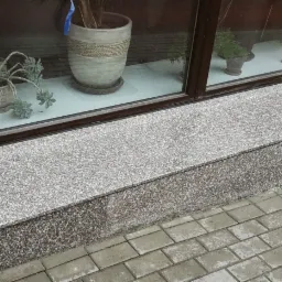 Zewnętrzny parapet z szarego, drobnoziarnistego konglomeratu, widoczny z poziomu chodnika wyłożonego kostką brukową, z roślinami doniczkowymi w tle za szybą okienną.