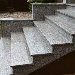 Zewnętrzne schody z szarego, ziarnistego kamienia prowadzące do budynku, z widocznym fragmentem kostki brukowej i zieleni w tle.