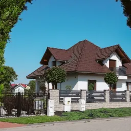 Biały dom jednorodzinny z brązowym dachem, widoczny przez ramę z zielonych krzewów, z szarym ogrodzeniem z elementami kamiennymi i ozdobnymi drzewkami.