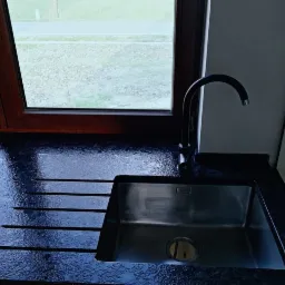 Czarny, błyszczący blat z kamienia z ociekaczem i wpuszczonym, prostokątnym zlewem ze stali nierdzewnej, bateria w kolorze czarnym, widok na okno w drewnianej ramie.
