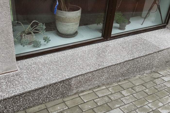 Zewnętrzny parapet z szarego, drobnoziarnistego konglomeratu, widoczny z poziomu chodnika wyłożonego kostką brukową, z roślinami doniczkowymi w tle za szybą okienną.