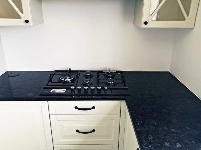 Czarny, błyszczący blat kuchenny z wbudowaną czarną płytą gazową, kremowe szafki z czarnymi uchwytami, białe ściany, widok z góry pod kątem.