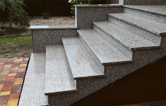 Zewnętrzne schody z szarego, ziarnistego kamienia prowadzące do budynku, z widocznym fragmentem kostki brukowej i zieleni w tle.