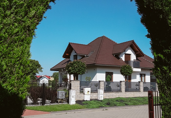 Biały dom jednorodzinny z brązowym dachem, widoczny przez ramę z zielonych krzewów, z szarym ogrodzeniem z elementami kamiennymi i ozdobnymi drzewkami.