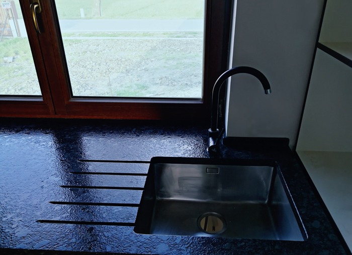 Czarny, błyszczący blat z kamienia z ociekaczem i wpuszczonym, prostokątnym zlewem ze stali nierdzewnej, bateria w kolorze czarnym, widok na okno w drewnianej ramie.