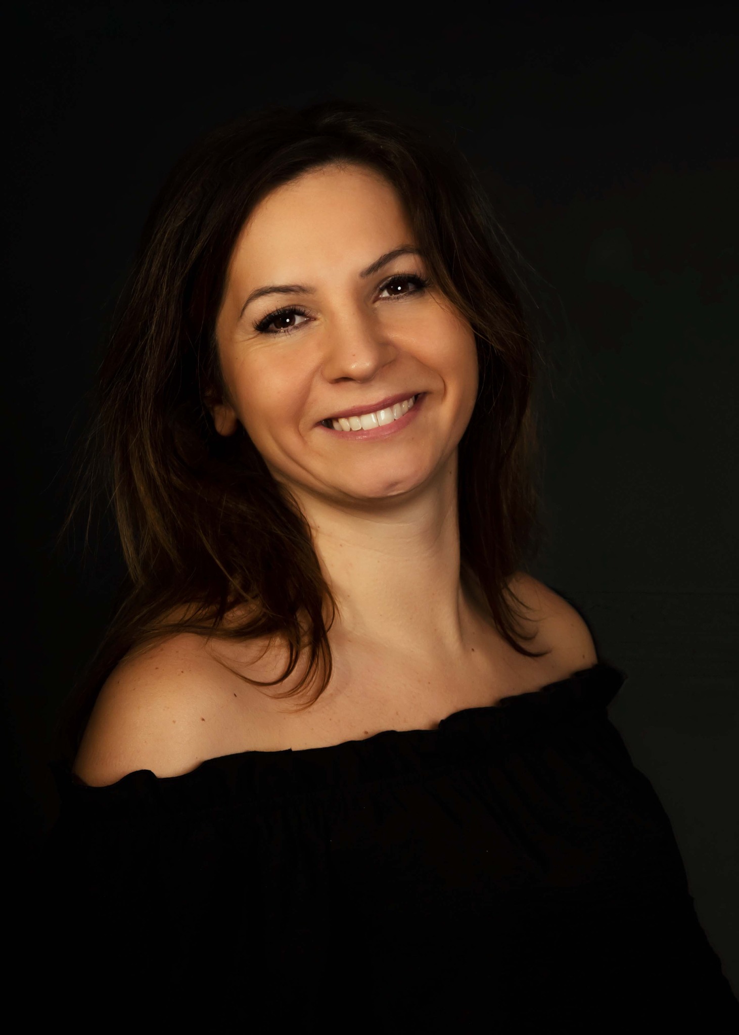 Portret uśmiechniętej brunetki w czarnej bluzce z odkrytymi ramionami na ciemnym tle.