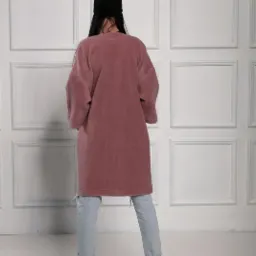 Kobieta tyłem w luźnym, długim swetrze w odcieniu pudrowego różu, czarnej czapce i jasnych dżinsach z przetarciami, na tle białej ściany z dekoracyjnymi panelami.