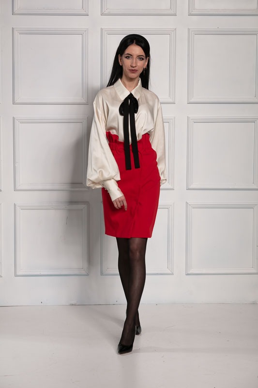 Kobieta w kremowej bluzce z czarną kokardą i czerwonej spódnicy ołówkowej, pozująca na tle białej ściany z panelami.