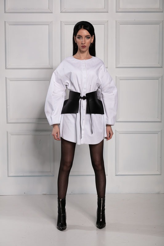 Kobieta w białej koszuli oversize z czarnym, skórzanym pasem gorsetowym, czarnych rajstopach i botkach na obcasie, pozująca na tle białych drzwi panelowych.
