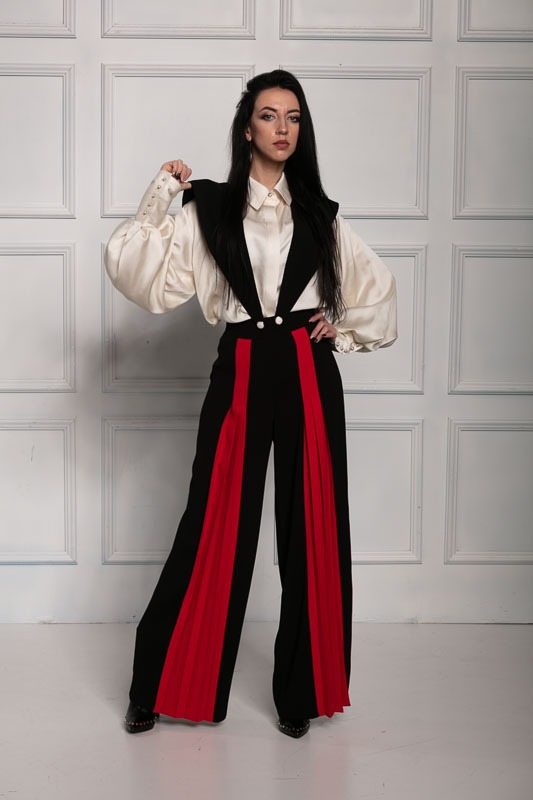 Kobieta w szerokich, czarno-czerwonych spodniach z szelkami i kremowej bluzce z bufiastymi rękawami, pozująca na tle ściany z kasetonami.