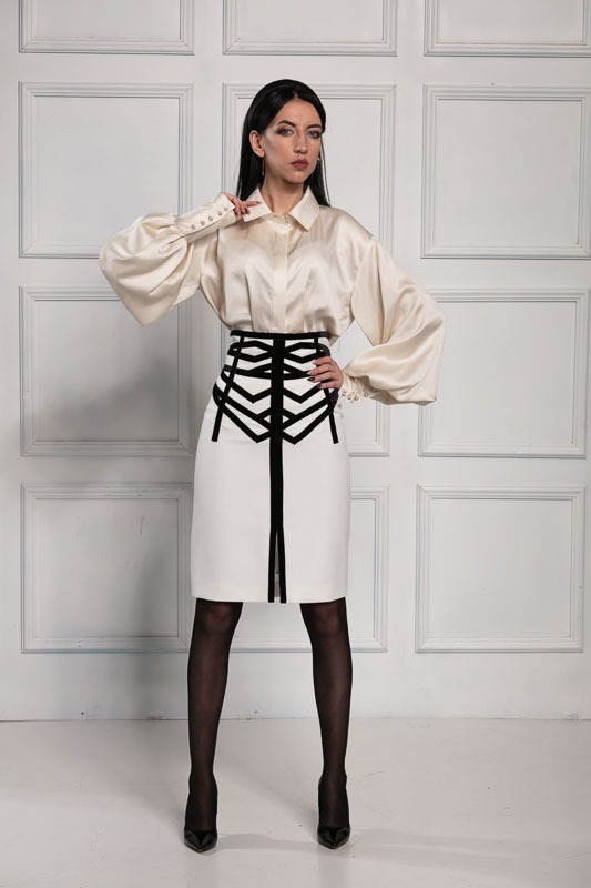 Kobieta w satynowej bluzce z bufiastymi rękawami i białej spódnicy z geometrycznym czarnym wzorem, pozująca na tle ściany z dekoracyjnymi panelami.