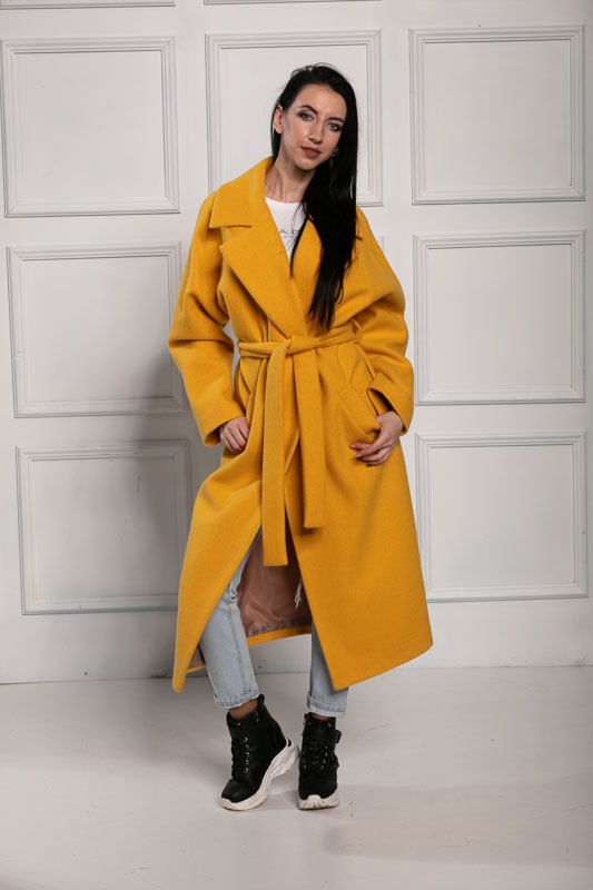 Kobieta w długim, żółtym płaszczu z paskiem, dżinsach i czarnych butach na białym tle z dekoracyjnymi panelami ściennymi.