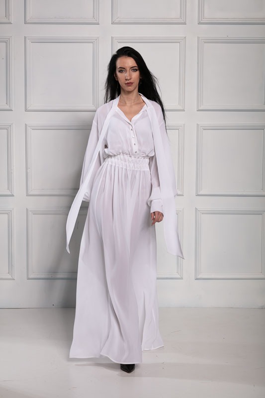 Kobieta w długiej, białej, zwiewnej sukience z bluzką koszulową, stojąca na tle białej ściany z dekoracyjnymi panelami.