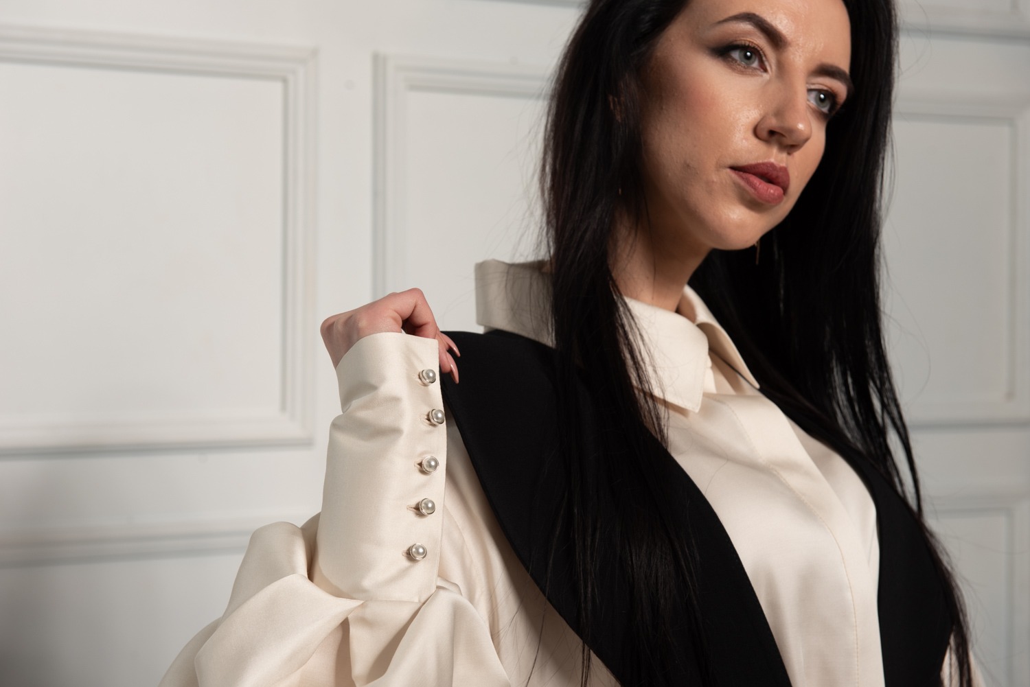 Kobieta w kremowej bluzce z ozdobnymi perłowymi guzikami na mankietach i czarnej kamizelce, pozująca na tle białej ściany z panelami.