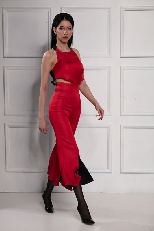 Kobieta prezentuje czerwoną, dwuczęściową kreację z wycięciem w talii i rozcięciami na nogawkach spodni, uzupełnioną czarnymi rajstopami i szpilkami na tle białych drzwi.