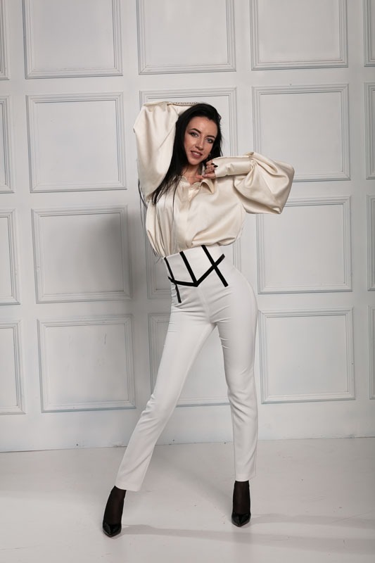 Kobieta w białych spodniach z czarnym wzorem na pasie i kremowej bluzce z bufiastymi rękawami, pozująca na tle białej ściany z panelami.