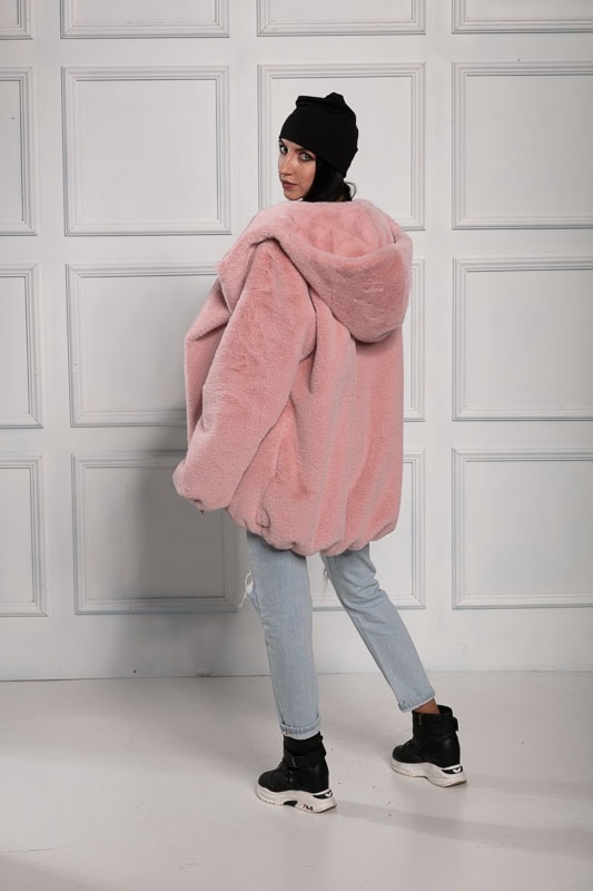 Kobieta w czarnej czapce i różowym, futrzanym płaszczu z kapturem, odwrócona plecami, prezentuje stylizację z jasnymi jeansami i czarnymi butami na platformie.