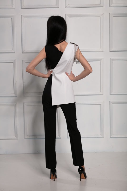 Kobieta w czarno-białym, asymetrycznym stroju z dekoracyjnymi łańcuszkami na ramieniu, pozująca tyłem na tle białej ściany z panelami.