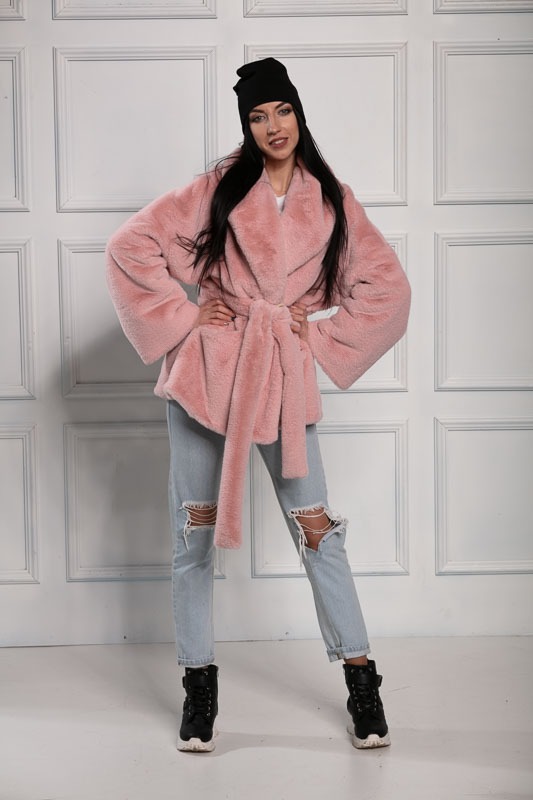 Kobieta w różowym, futrzanym płaszczu z wiązaniem w pasie, czarnej czapce beanie i podartych jeansach, pozująca na tle białej ściany z panelami.