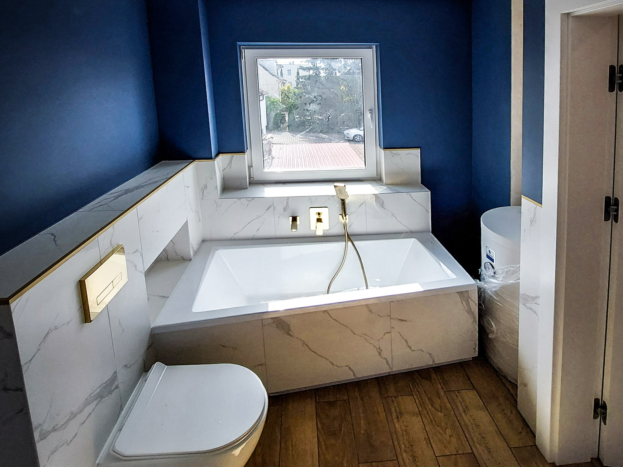 Nowoczesna łazienka z głęboką wanną obudowaną marmurowymi płytami z delikatnym złotym akcentem, widok na okno z zielenią na zewnątrz, toaleta w rogu i drewniana podłoga