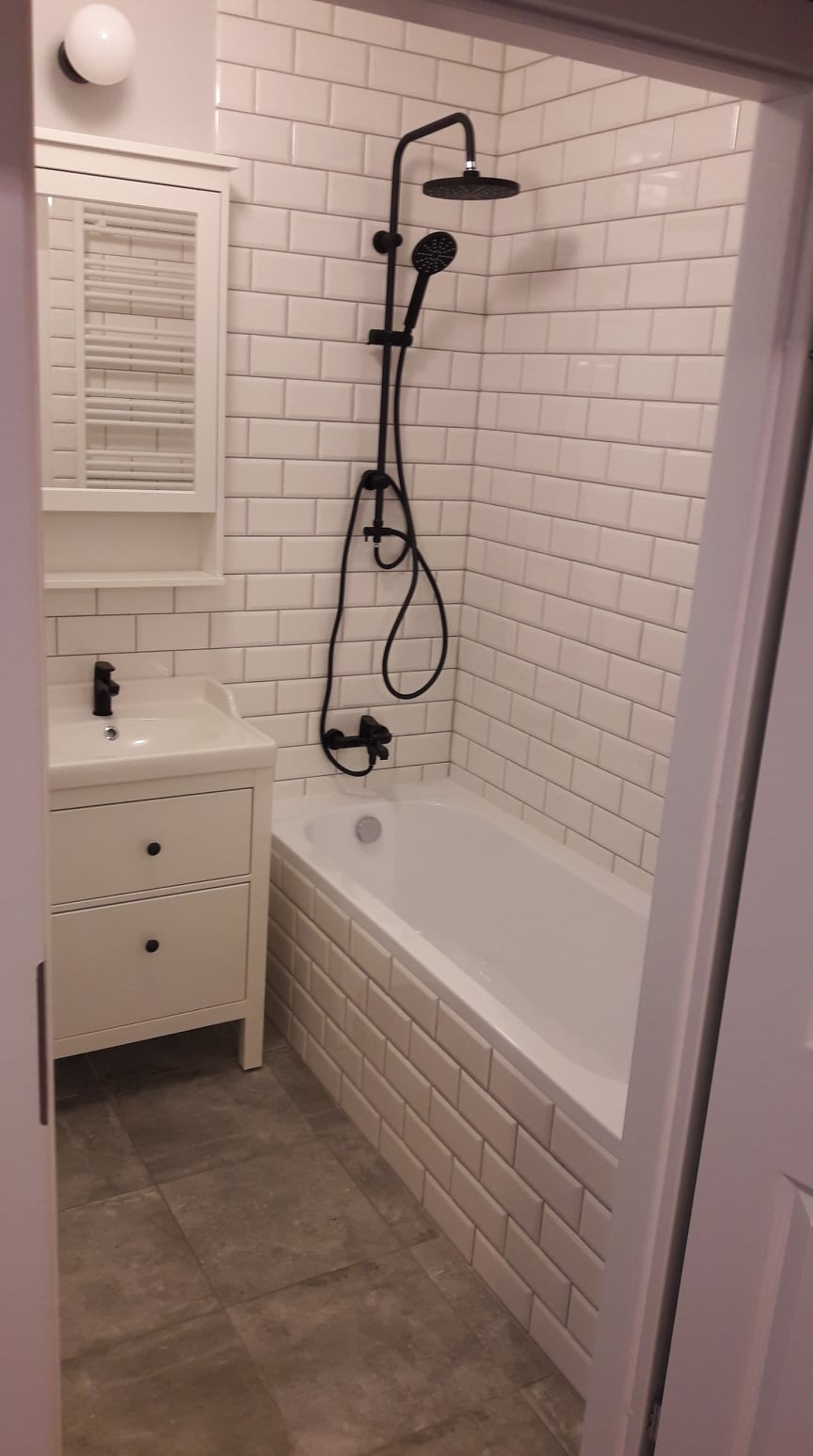 Wykończona łazienka z białą armaturą, czarną baterią prysznicową i umywalką z szafką, wyłożona białymi płytkami typu metro i szarą podłogą.