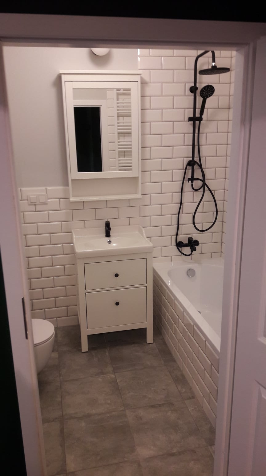 Nowoczesna łazienka z białymi płytkami typu metro, białą szafką z umywalką i czarną armaturą, widok zza otwartych drzwi.