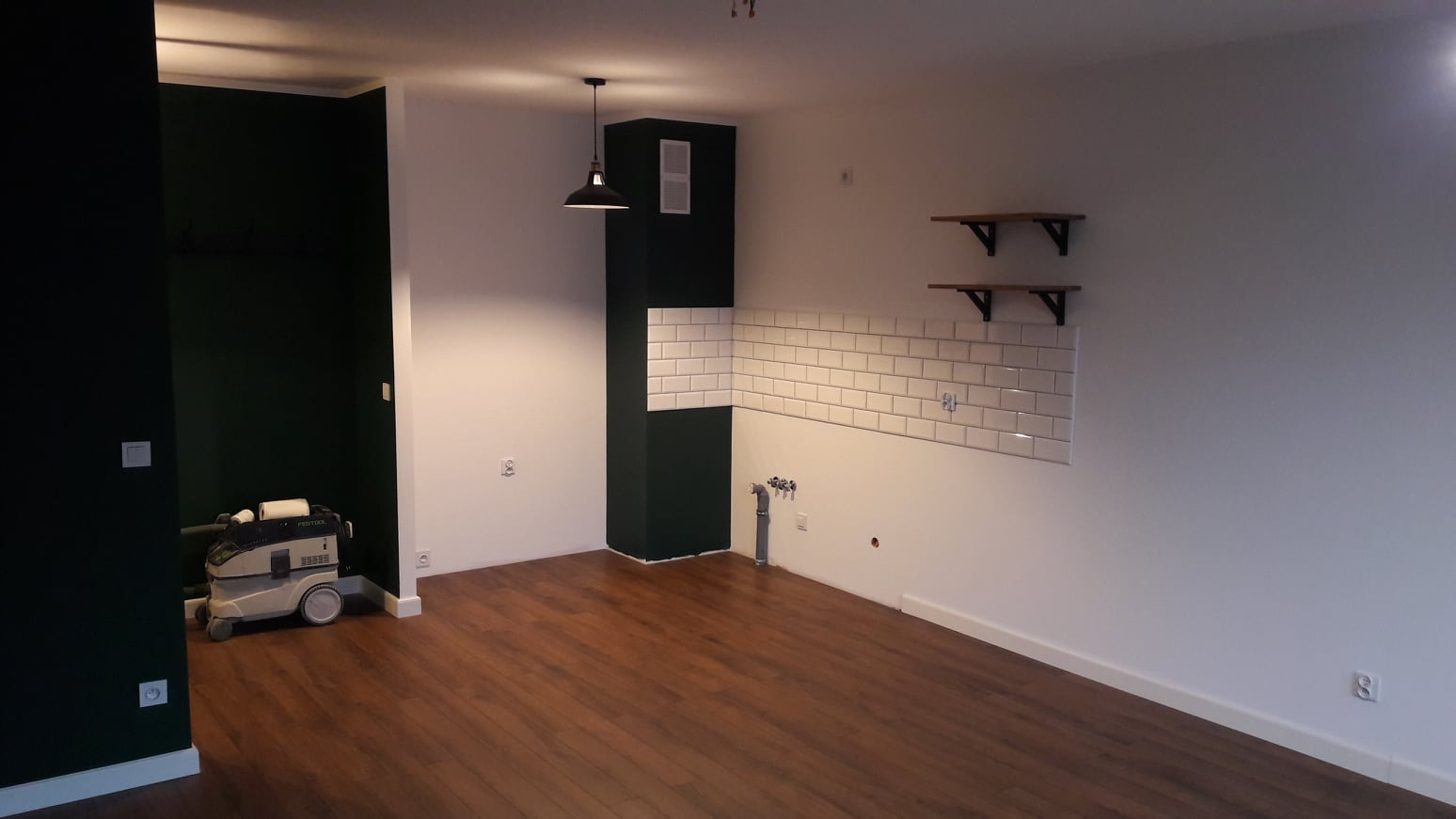 Wnętrze po remoncie: ciemnozielona ściana, ułożona glazura w stylu metro, drewniana podłoga, wiszące półki i odkurzacz przemysłowy Festool.