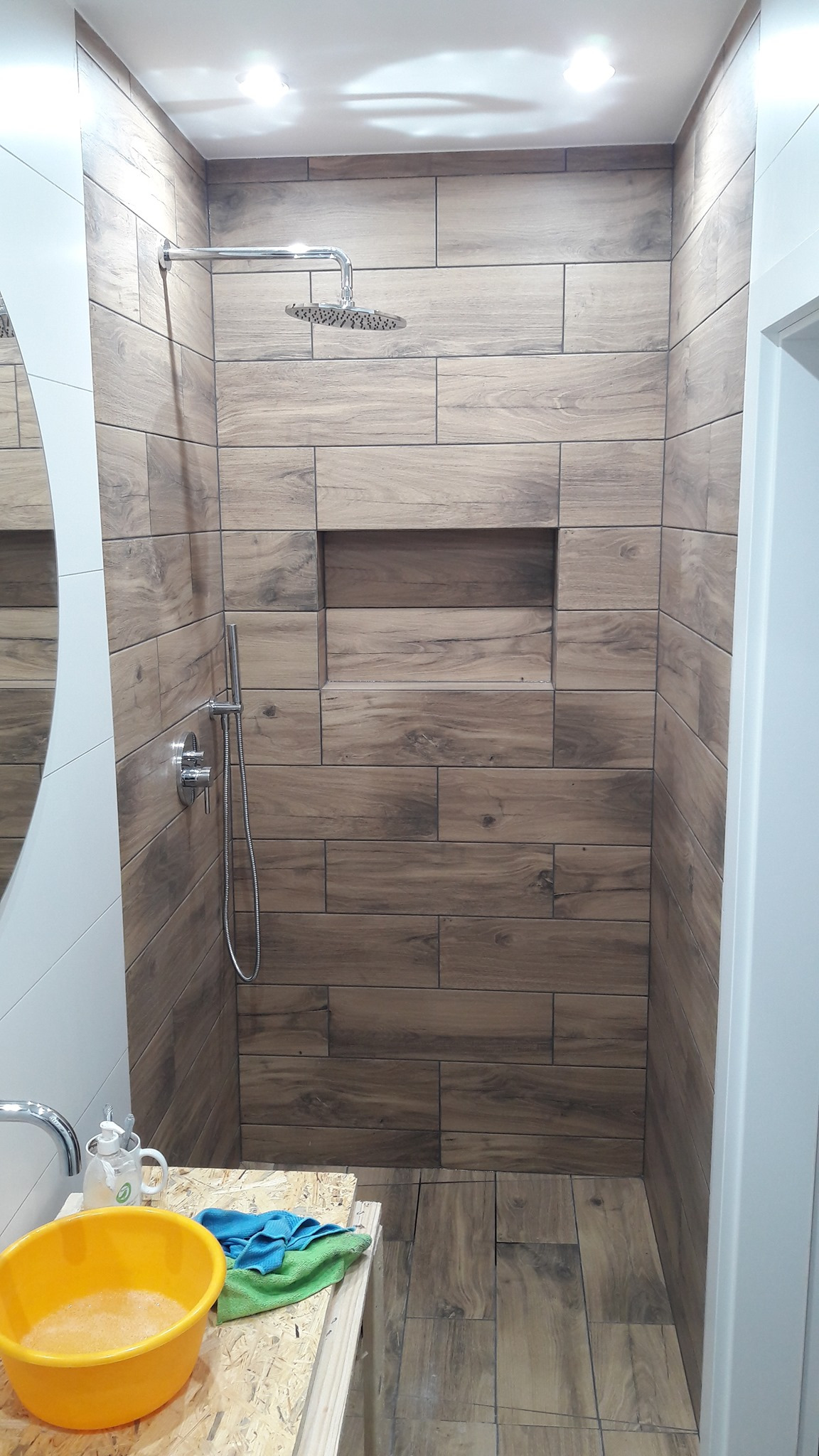 Kabina prysznicowa z płytkami imitującymi drewno, wnęka w ścianie, bateria i deszczownica, widok na żółtą miskę z wodą i narzędzia pracy w Dobrej.