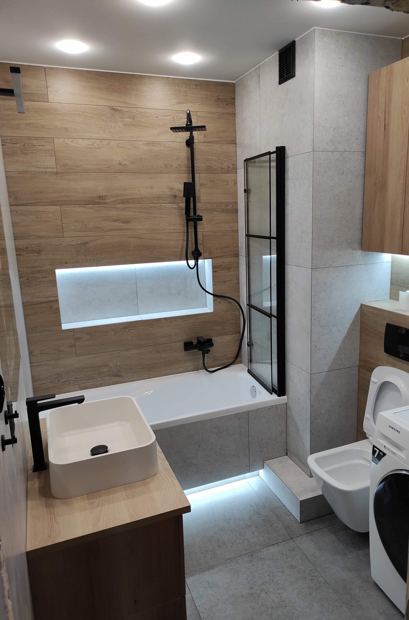 Nowoczesna łazienka z drewnopodobnymi płytkami, kabiną nawannową z czarnymi profilami, prostokątną umywalką nablatową i podświetleniem LED pod wanną oraz w niszy ściennej.