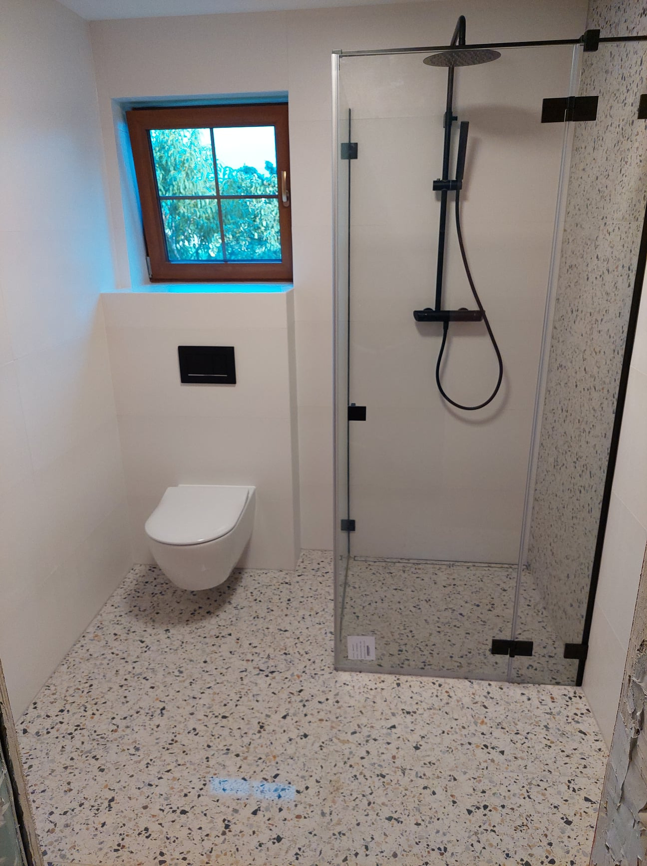 Nowoczesna łazienka z kabiną prysznicową typu walk-in, podwieszaną toaletą i podłogą z lastryko, oświetlona przez okno z drewnianą ramą.