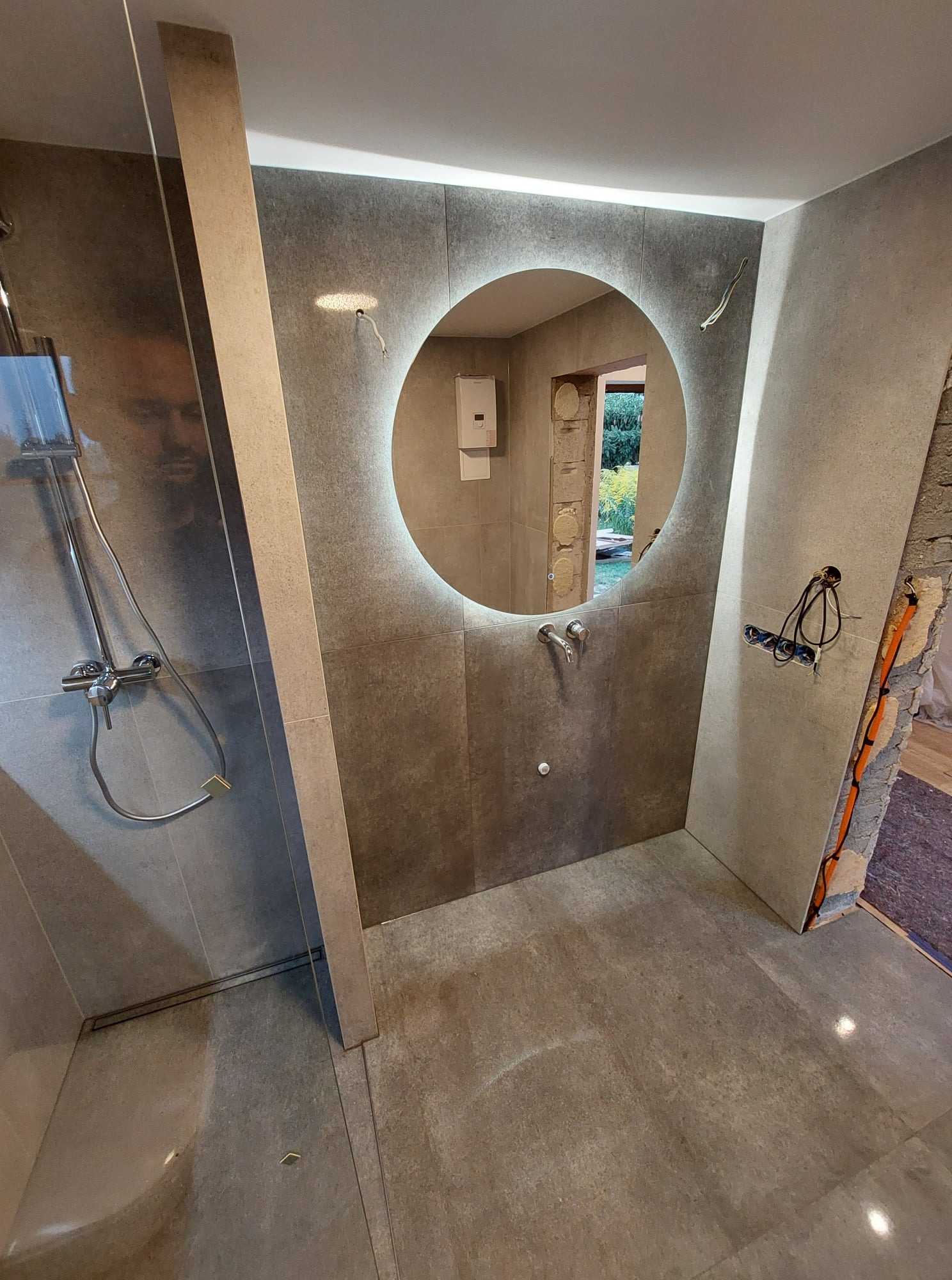 Wnętrze łazienki w trakcie prac wykończeniowych: szara glazura ścienna i podłogowa, okrągłe lustro z podświetleniem LED, widoczne przewody elektryczne, kabina prysznicowa ze szklaną ścianką.