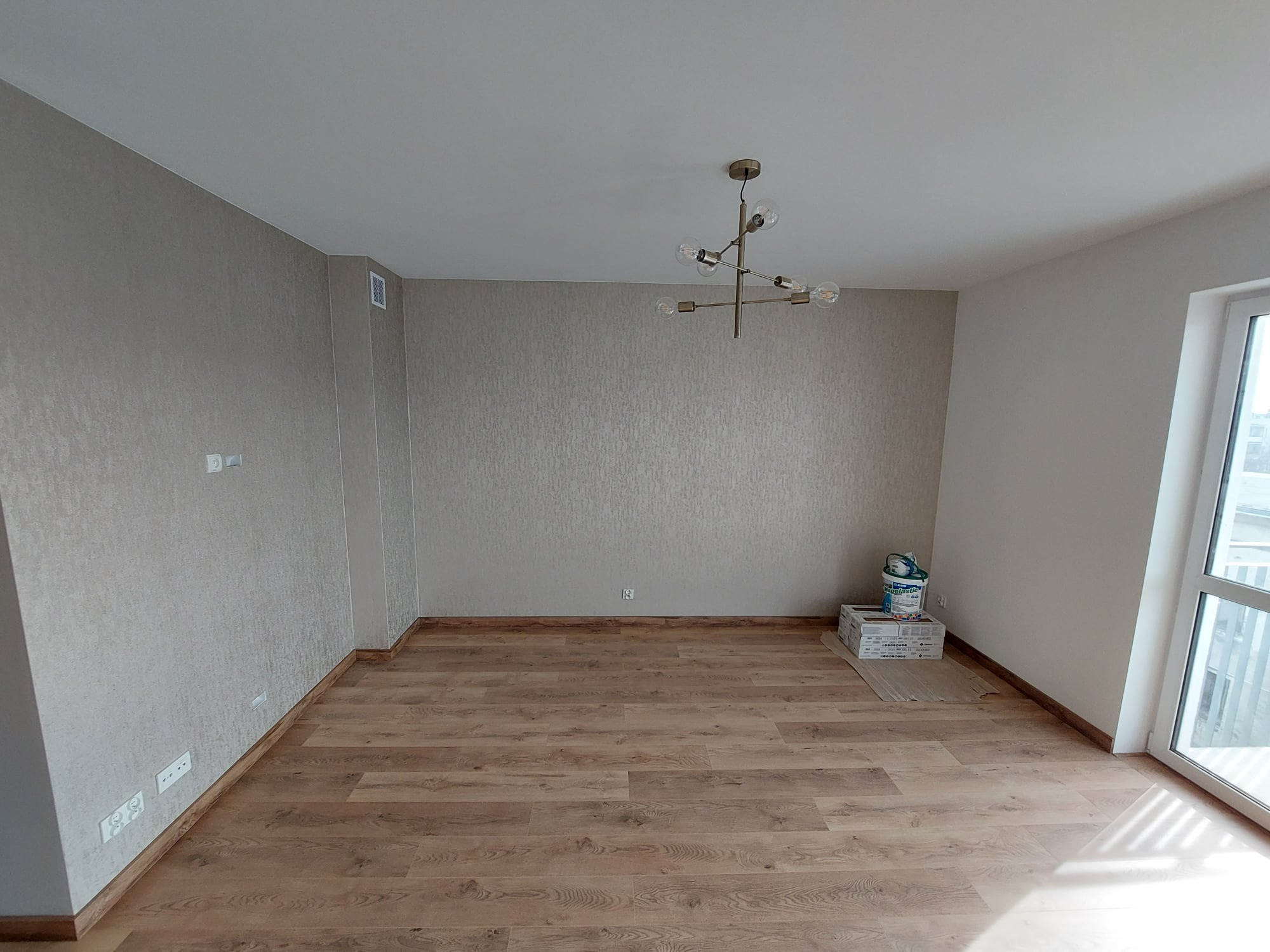Pusty pokój po remoncie z panelami podłogowymi w odcieniach brązu, beżowe ściany z delikatną fakturą, nowoczesny żyrandol z sześcioma żarówkami, kartony i puszka z materiałami budowlanymi w rogu...